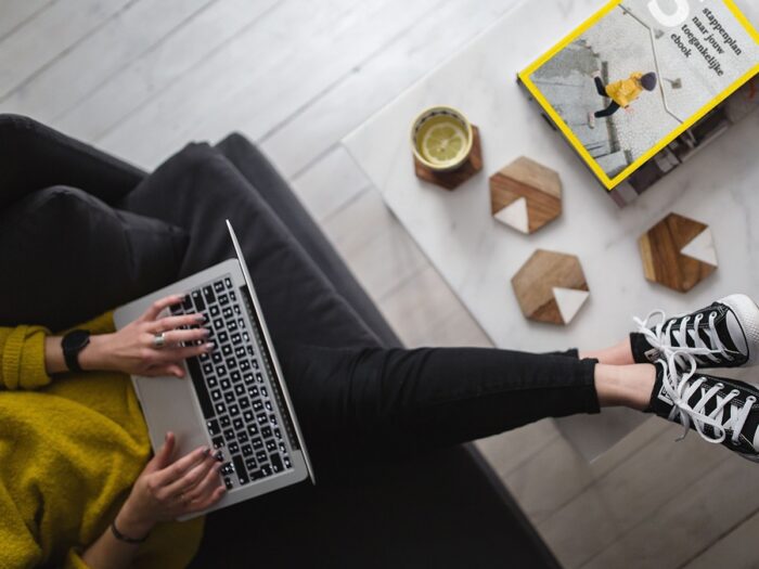 Vrouw zit met laptop op de bank. Op tafel een stapeltje boeken.